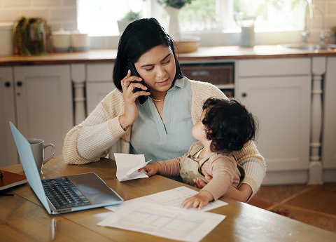 Mother holding phone up to ear and looking at child while both are sitting on a desk with a laptop
