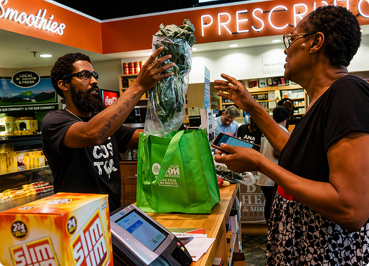 At a grocery store checkout counter, a staff member bags fresh leafy greens for a customer, highlighting access to fresh produce in a retail food setting
