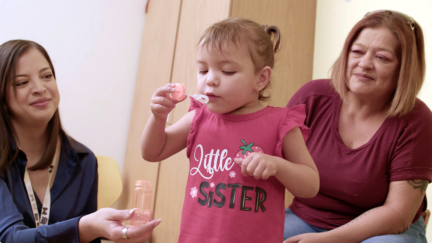 A young child blows bubbles while two seated adults observe.