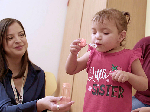A young child blows bubbles while two seated adults observe.