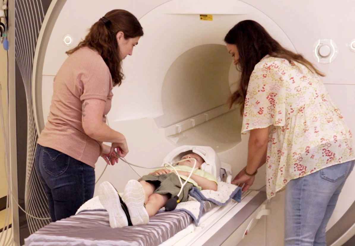 A child lies on the table of a medical imaging scanner while two adults position the child and equipment for the scan.