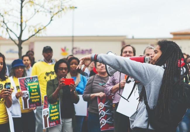 A public gathering outside a grocery store, where a speaker addresses community members with a megaphone while attendees hold signs advocating for healthy food 
