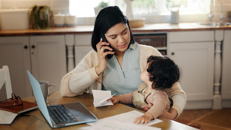 Mother holding phone up to ear and looking at child while both are sitting on a desk with a laptop on it
