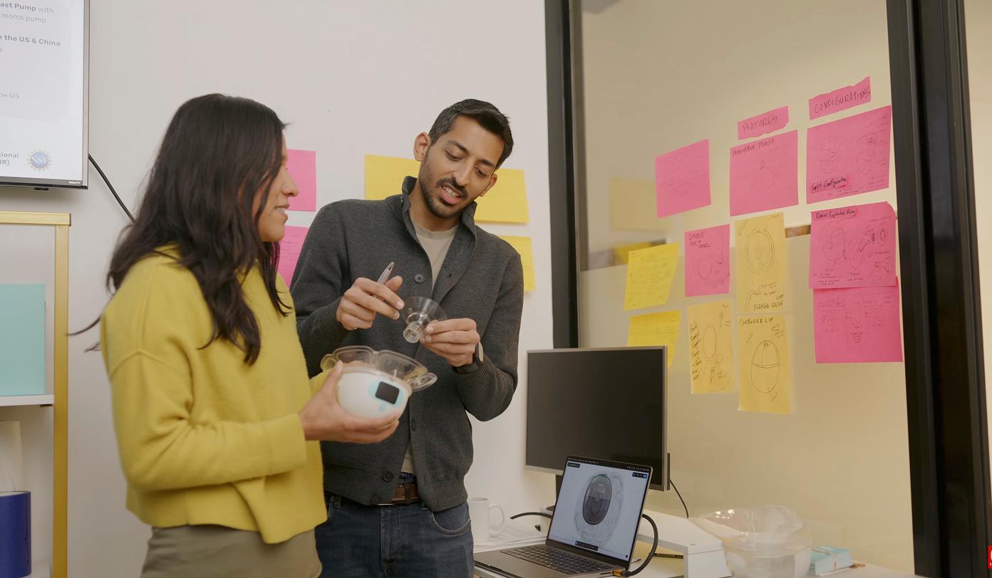 Two scientist examining a small device in an office with sticky notes on the wall and a laptop on a desk