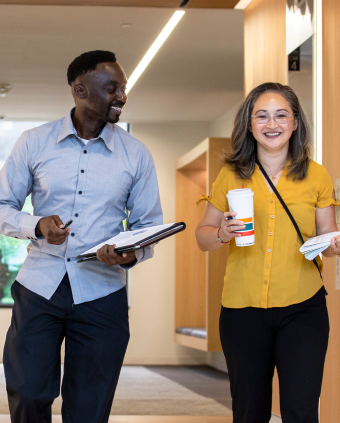 Two IQ Solutions employees walking down hall and smiling one person is holding a clipboard and other person is holding coffee cup